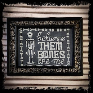 May include: A black framed cross-stitch with a white skeleton and the text "Believe I Them Bones Are Me".