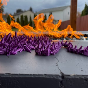 Puede incluir: Una figura de dragón de color púrpura, impresa en 3D, con púas. El dragón está segmentado y tiene una cola larga. La figura está sentada sobre una superficie gris.