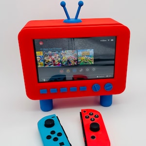 May include: A red and blue retro-style television console with a Nintendo Switch screen displaying the game Mario Kart 8 Deluxe. Two Nintendo Switch Joy-Con controllers are shown, one blue and one red.