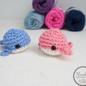 Puede incluir: Dos juguetes de ballena de ganchillo, uno azul claro y otro rosa, cada uno con vientre blanco y ojos negros. Est&aacute;n sobre una superficie blanca, con ovillos de lana en morado, azul, rosa y marino en el fondo. Juguetes hechos a mano.