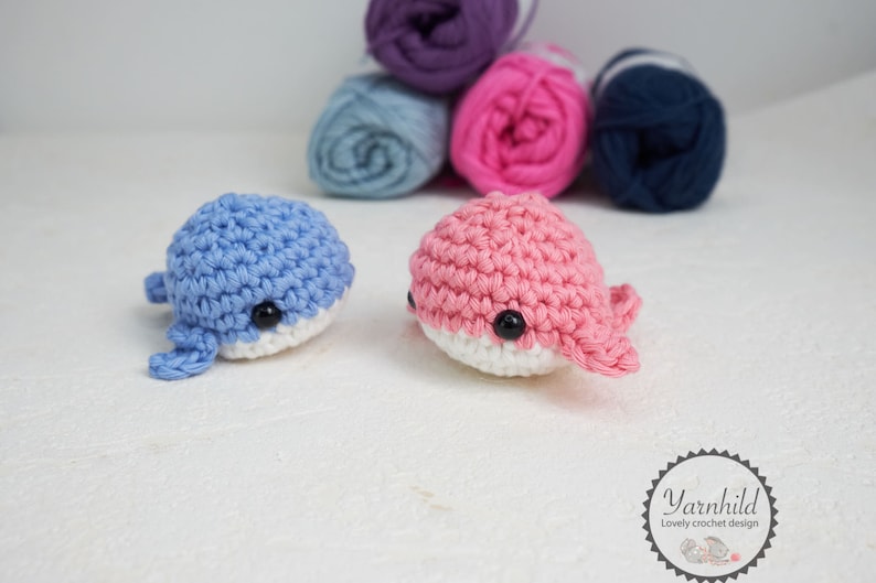 Puede incluir: Dos juguetes de ballena de ganchillo, uno azul claro y otro rosa, cada uno con vientre blanco y ojos negros. Est&aacute;n sobre una superficie blanca, con ovillos de lana en morado, azul, rosa y marino en el fondo. Juguetes hechos a mano.
