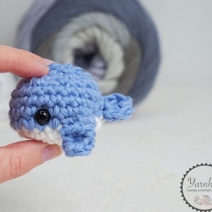 Puede incluir: Una peque&ntilde;a ballena azul tejida a ganchillo con detalles blancos y un ojo negro. El juguete hecho a mano se sostiene frente a un fondo borroso de hilo. La ballena es un art&iacute;culo lindo y artesanal.