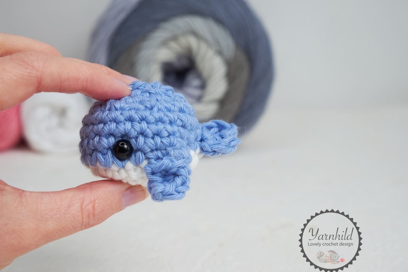 Puede incluir: Una peque&ntilde;a ballena azul tejida a ganchillo con detalles blancos y un ojo negro. El juguete hecho a mano se sostiene frente a un fondo borroso de hilo. La ballena es un art&iacute;culo lindo y artesanal.