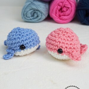 Puede incluir: Dos juguetes de ballena de ganchillo, uno azul y otro rosa, con vientres blancos y ojos negros. Las ballenas est&aacute;n hechas a mano y se encuentran frente a varios ovillos de lana en tonos morado, azul, rosa y azul marino. La imagen destaca la textura de la lana.