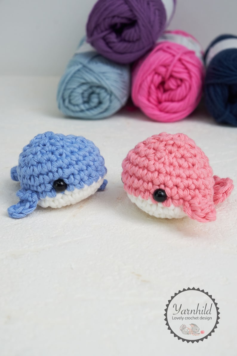 Puede incluir: Dos juguetes de ballena de ganchillo, uno azul y otro rosa, con vientres blancos y ojos negros. Las ballenas est&aacute;n hechas a mano y se encuentran frente a varios ovillos de lana en tonos morado, azul, rosa y azul marino. La imagen destaca la textura de la lana.