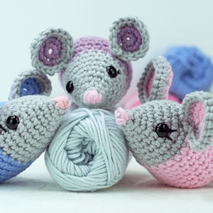 May include: Three crocheted gray mice with pink and blue accents. The mice are sitting on a white surface with a ball of light blue yarn in the center.