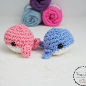 Puede incluir: Dos juguetes de ballena de ganchillo, uno rosa y otro azul, con vientres blancos y ojos negros. Las ballenas est&aacute;n hechas a mano y se encuentran sobre una superficie blanca. Ovillos de lana en morado, azul y rosa en el fondo.