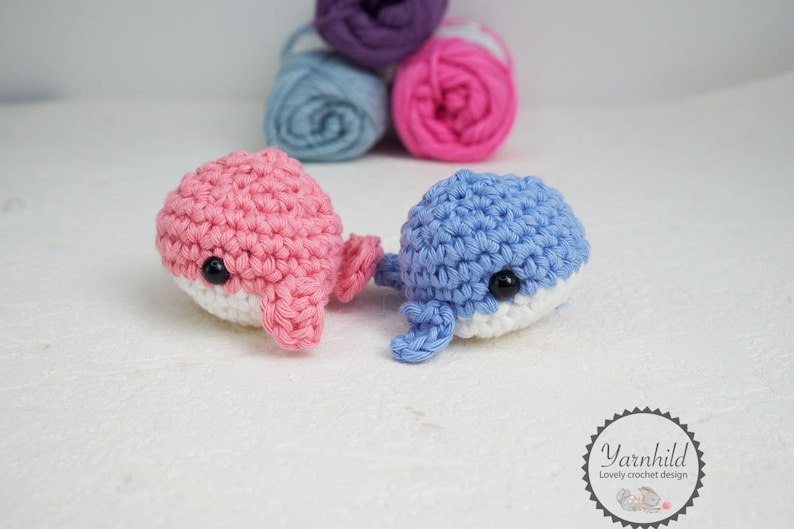 Puede incluir: Dos juguetes de ballena de ganchillo, uno rosa y otro azul, con vientres blancos y ojos negros. Las ballenas est&aacute;n hechas a mano y se encuentran sobre una superficie blanca. Ovillos de lana en morado, azul y rosa en el fondo.