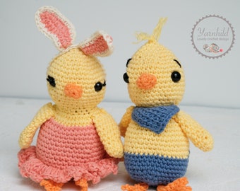 May include: Two crocheted yellow chicks, one wearing a pink bunny ear headband and a pink dress, the other wearing a blue scarf and blue pants.  The chicks have black eyes and orange beaks.  The text "Yarnhild Lovely crochet designs" is visible in the upper right corner of the image.