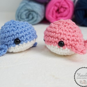 Puede incluir: Dos juguetes de ballena de ganchillo, uno azul y otro rosa, con vientres blancos y ojos de bot&oacute;n negros. Las ballenas est&aacute;n hechas a mano y se encuentran sobre una superficie blanca. Ovillos de lana en azul, rosa y azul marino en el fondo. El logo "Yarnhild" es visible.