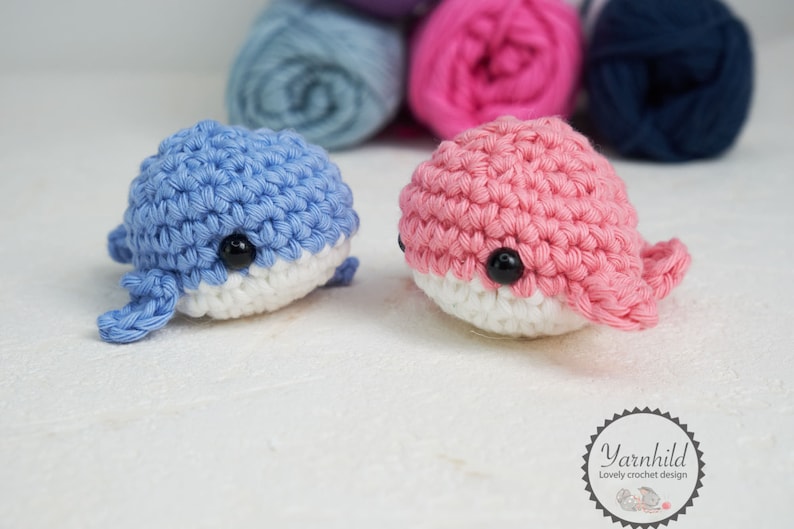 Puede incluir: Dos juguetes de ballena de ganchillo, uno azul y otro rosa, con vientres blancos y ojos de bot&oacute;n negros. Las ballenas est&aacute;n hechas a mano y se encuentran sobre una superficie blanca. Ovillos de lana en azul, rosa y azul marino en el fondo. El logo "Yarnhild" es visible.