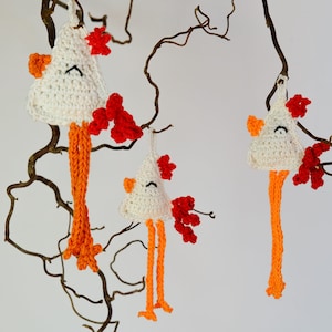 Puede incluir: Tres adornos de gallina hechos a mano con ganchillo colgando de una rama. Cada gallina tiene una cabeza blanca, pico y patas naranjas y una cresta roja. Las gallinas están suspendidas sobre un fondo blanco.