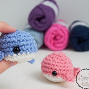 May include: Two crocheted whale toys, one light blue and one pink, each with a white belly and black eyes. The blue whale is held by a hand. Behind them are several balls of yarn in shades of purple, pink, and blue.