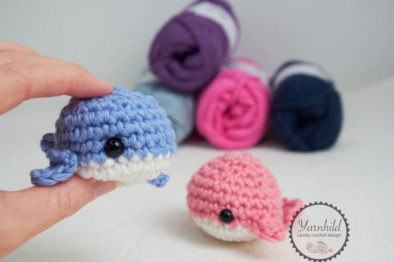 Puede incluir: Dos juguetes de ballena de ganchillo, uno azul claro y otro rosa, cada uno con vientre blanco y ojos negros. La ballena azul est&aacute; sujeta por una mano. Detr&aacute;s hay varios ovillos de lana en tonos morados, rosas y azules.
