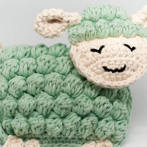 May include: A handmade crochet sheep toy in soft green and cream colors. The sheep has a textured body, cream-colored ears, and a smiling face with black embroidered details. This adorable stuffed animal is perfect for a nursery or as a decorative item.