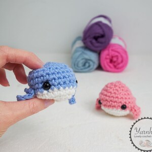 Puede incluir: Una mano sostiene una peque&ntilde;a ballena azul tejida a ganchillo con detalles blancos y ojos negros. Tambi&eacute;n se ve una ballena rosa tejida a ganchillo. Detr&aacute;s hay ovillos de lana morada, azul y rosa. Imagen de art&iacute;culos de ganchillo hechos a mano.