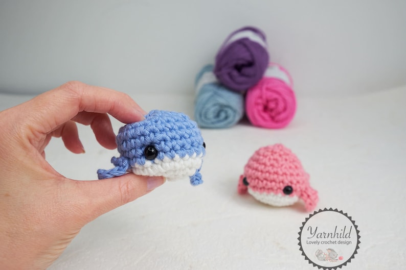 Puede incluir: Una mano sostiene una peque&ntilde;a ballena azul tejida a ganchillo con detalles blancos y ojos negros. Tambi&eacute;n se ve una ballena rosa tejida a ganchillo. Detr&aacute;s hay ovillos de lana morada, azul y rosa. Imagen de art&iacute;culos de ganchillo hechos a mano.