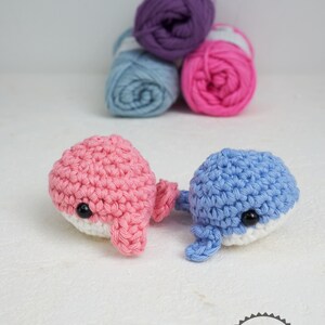 Puede incluir: Dos juguetes de ballena de ganchillo, uno rosa y otro azul, con vientres blancos y ojos negros. Detr&aacute;s, tres ovillos de lana morada, azul y rosa. Los juguetes est&aacute;n hechos a mano, probablemente para ni&ntilde;os.