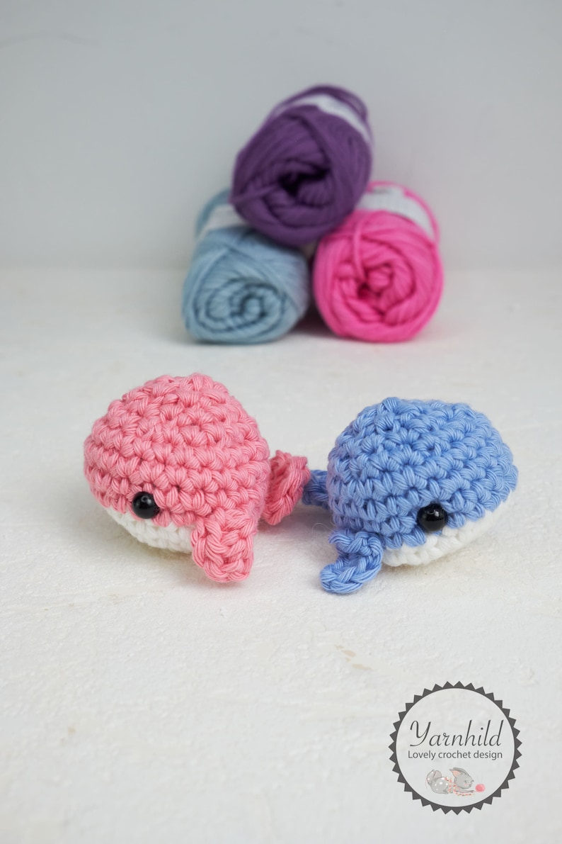 Puede incluir: Dos juguetes de ballena de ganchillo, uno rosa y otro azul, con vientres blancos y ojos negros. Detr&aacute;s, tres ovillos de lana morada, azul y rosa. Los juguetes est&aacute;n hechos a mano, probablemente para ni&ntilde;os.