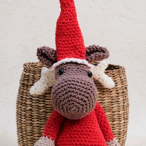 Peut inclure: Un élan en crochet portant un bonnet de Père Noël rouge avec un pompon blanc. L'élan porte un pull rouge avec des bordures blanches et a des bois bruns.