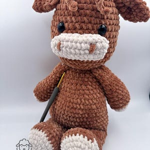 Puede incluir: Una vaca de peluche de crochet marrón con detalles blancos en la cara y los pies. La vaca está sentada con un gancho de crochet delante de ella.