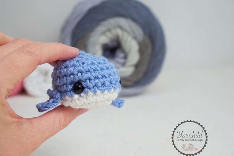 Puede incluir: Una peque&ntilde;a ballena azul y blanca tejida a ganchillo con ojos negros. La ballena se sostiene en una mano, con una madeja de hilo en el fondo. El hilo es un degradado de azul, gris y blanco. El objeto es hecho a mano.