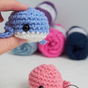 Puede incluir: Juguetes de ballena de ganchillo hechos a mano en azul y rosa, con vientres blancos y ojos negros. La imagen tambi&eacute;n muestra varias madejas de lana en varios colores, lo que sugiere un tema de artesan&iacute;a. Los juguetes son peque&ntilde;os y detallados.