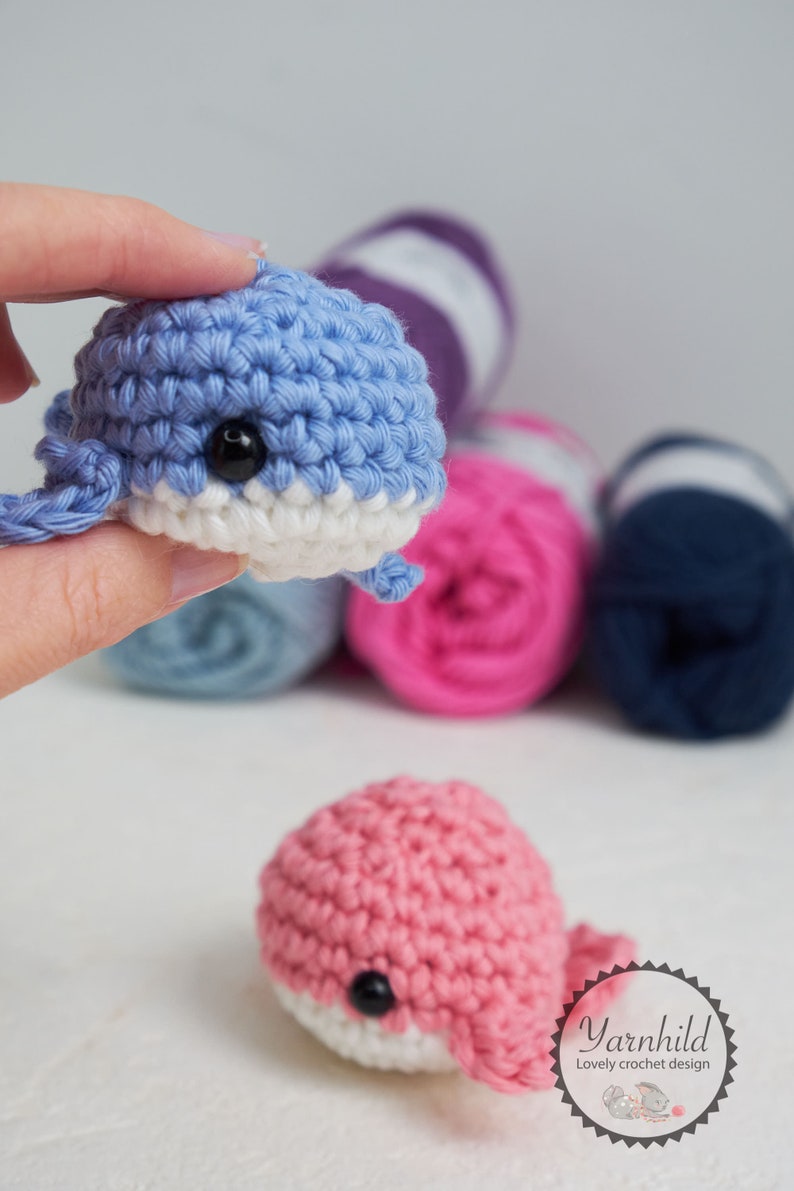 Puede incluir: Juguetes de ballena de ganchillo hechos a mano en azul y rosa, con vientres blancos y ojos negros. La imagen tambi&eacute;n muestra varias madejas de lana en varios colores, lo que sugiere un tema de artesan&iacute;a. Los juguetes son peque&ntilde;os y detallados.