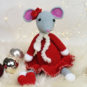 May include: A crocheted gray mouse wearing a red dress with white trim and a white fur scarf. The mouse is holding a red heart in its paws and is sitting on a white fluffy surface.