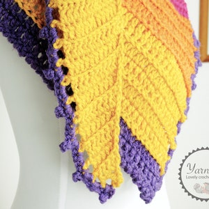 May include: Close-up of a crocheted shawl with a leaf pattern in yellow, orange, and purple yarn. The shawl is crocheted in a textured stitch and has a scalloped edge.