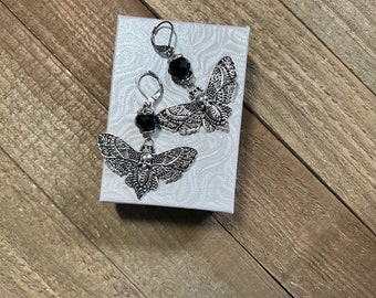May include: A pair of silver moth earrings with black glass beads. The earrings are displayed on a white box with a textured surface.
