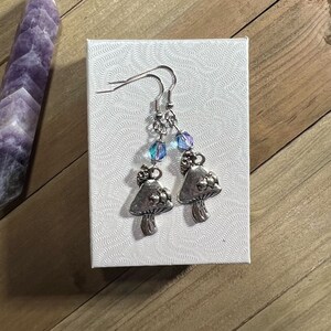 Puede incluir: Un par de pendientes de plata con forma de hongo con cuentas azules y moradas iridiscentes que cuelgan de las tapas de los hongos.