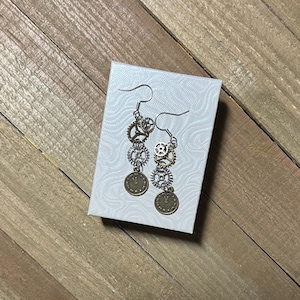 May include: A pair of silver and bronze earrings with a clock face and gears hanging from the bottom. The earrings are displayed on a white box with a textured surface.