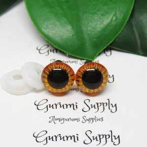 May include: Two brown safety eyes with black pupils and a gold rim. The eyes are on a white surface with a white plastic washer.