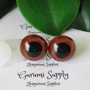 Puede incluir: Dos ojos de seguridad de pl&aacute;stico marr&oacute;n con pupilas negras, utilizados para amigurumi u otras manualidades. Los ojos est&aacute;n sobre una superficie blanca con una arandela blanca.