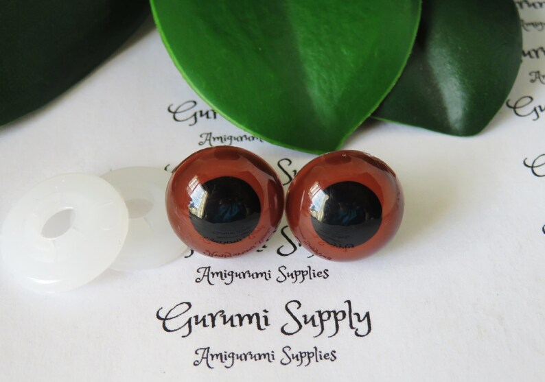 Puede incluir: Dos ojos de seguridad de pl&aacute;stico marr&oacute;n con pupilas negras, utilizados para amigurumi u otras manualidades. Los ojos est&aacute;n sobre una superficie blanca con una arandela blanca.