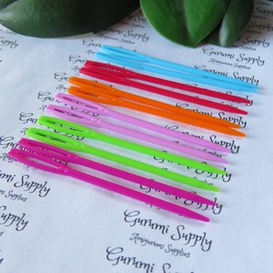 May include: A set of ten colorful plastic crochet hooks, arranged in a rainbow pattern. The hooks are labeled "Gurumi Supply" and are designed for amigurumi projects.