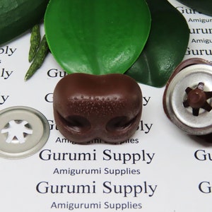 May include: Close-up of brown amigurumi nose and safety eyes with silver washers. The nose is a rounded shape with two nostril indentations and small dot details. The safety eyes have a silver washer.