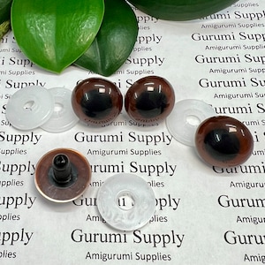 May include: Four brown safety eyes with black pupils and white plastic washers. The eyes are 12mm in diameter. The text "Gurumi Supply Amigurumi Supplies" is printed on the white surface.