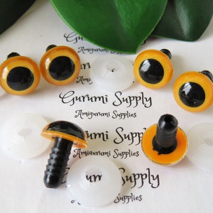 May include: A close-up of six orange and black safety eyes with black plastic backs. The eyes are arranged in a semi-circle on a white surface. The eyes are used for amigurumi and other crafts.
