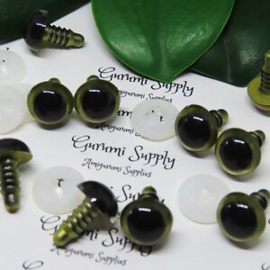 Puede incluir: Surtido de ojos de seguridad de pl&aacute;stico verde y negro con tornillos de metal dorado. Los ojos est&aacute;n esparcidos sobre una superficie blanca con el texto "Gurumi Supply Amigurumi Supplies" impreso en ella.