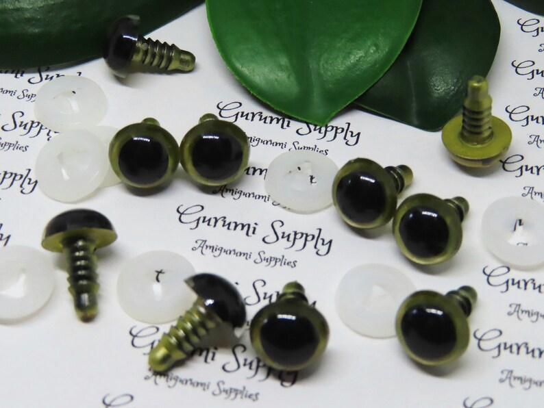Puede incluir: Surtido de ojos de seguridad de pl&aacute;stico verde y negro con tornillos de metal dorado. Los ojos est&aacute;n esparcidos sobre una superficie blanca con el texto "Gurumi Supply Amigurumi Supplies" impreso en ella.