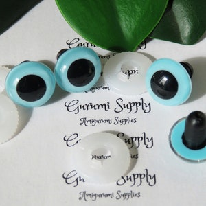 May include: Four blue plastic safety eyes with black pupils and white plastic washers. The eyes are for use in amigurumi projects. The text "Gurumi Supply Amigurumi Supplies" is visible on the white background.