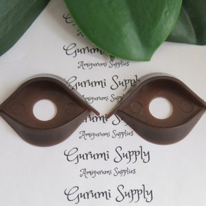 May include: Two brown plastic safety eyes for amigurumi projects. Each eye has a teardrop shape with a central hole and a small tab on each side. The eyes are designed for crafting and doll making.