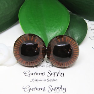 May include: Two brown glass safety eyes with black pupils and a brown iris. The eyes have a radial pattern around the pupil. The eyes are on a white surface with the text "Gurumi Supply Amigurumi Supplies" printed on the surface.