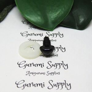 May include: A close-up of a black plastic safety eye with a white plastic washer. The eye is on a white surface with the text "Gurumi Supply" printed on it.