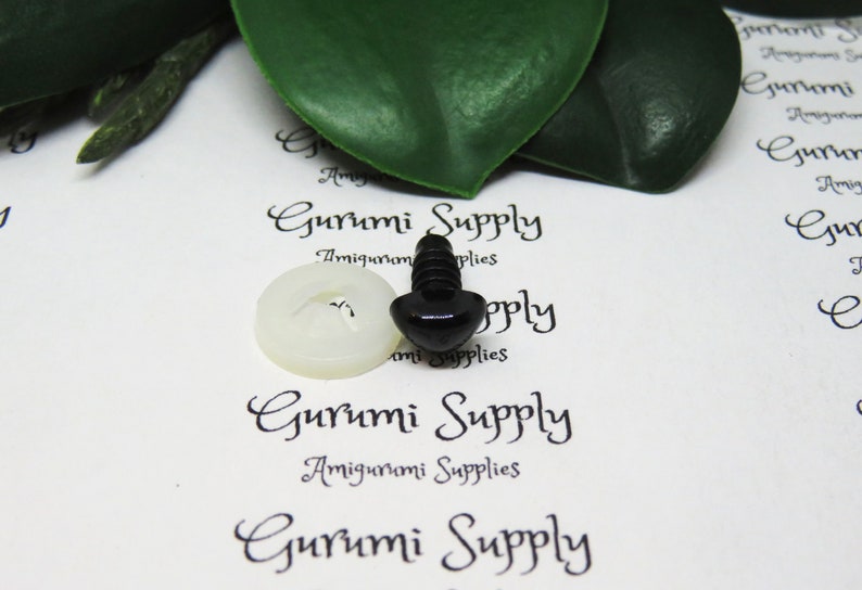 May include: A close-up of a black plastic safety eye with a white plastic washer. The eye is on a white surface with the text "Gurumi Supply" printed on it.