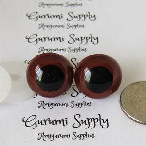 Puede incluir: Dos ojos de seguridad de pl&aacute;stico marr&oacute;n con pupilas negras, una arandela de pl&aacute;stico blanca y una moneda de 25 centavos de d&oacute;lar estadounidense para comparar el tama&ntilde;o. Gurumi Supply est&aacute; impreso en el fondo blanco.