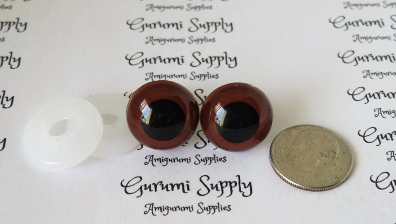 Puede incluir: Dos ojos de seguridad de pl&aacute;stico marr&oacute;n con pupilas negras, una arandela de pl&aacute;stico blanca y una moneda de 25 centavos de d&oacute;lar estadounidense para comparar el tama&ntilde;o. Gurumi Supply est&aacute; impreso en el fondo blanco.