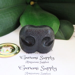 May include: Black plastic amigurumi nose with a textured surface. The nose has two nostrils and a small indentation in the center.